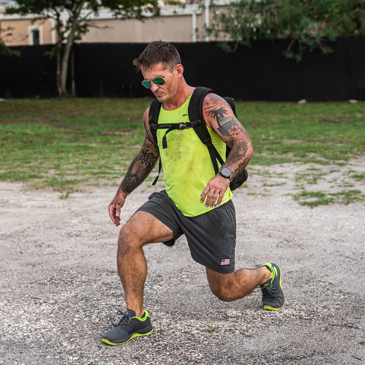 Fit man with tattoos in neon tank top and shorts performing lunge exercise outdoors wearing a backpack