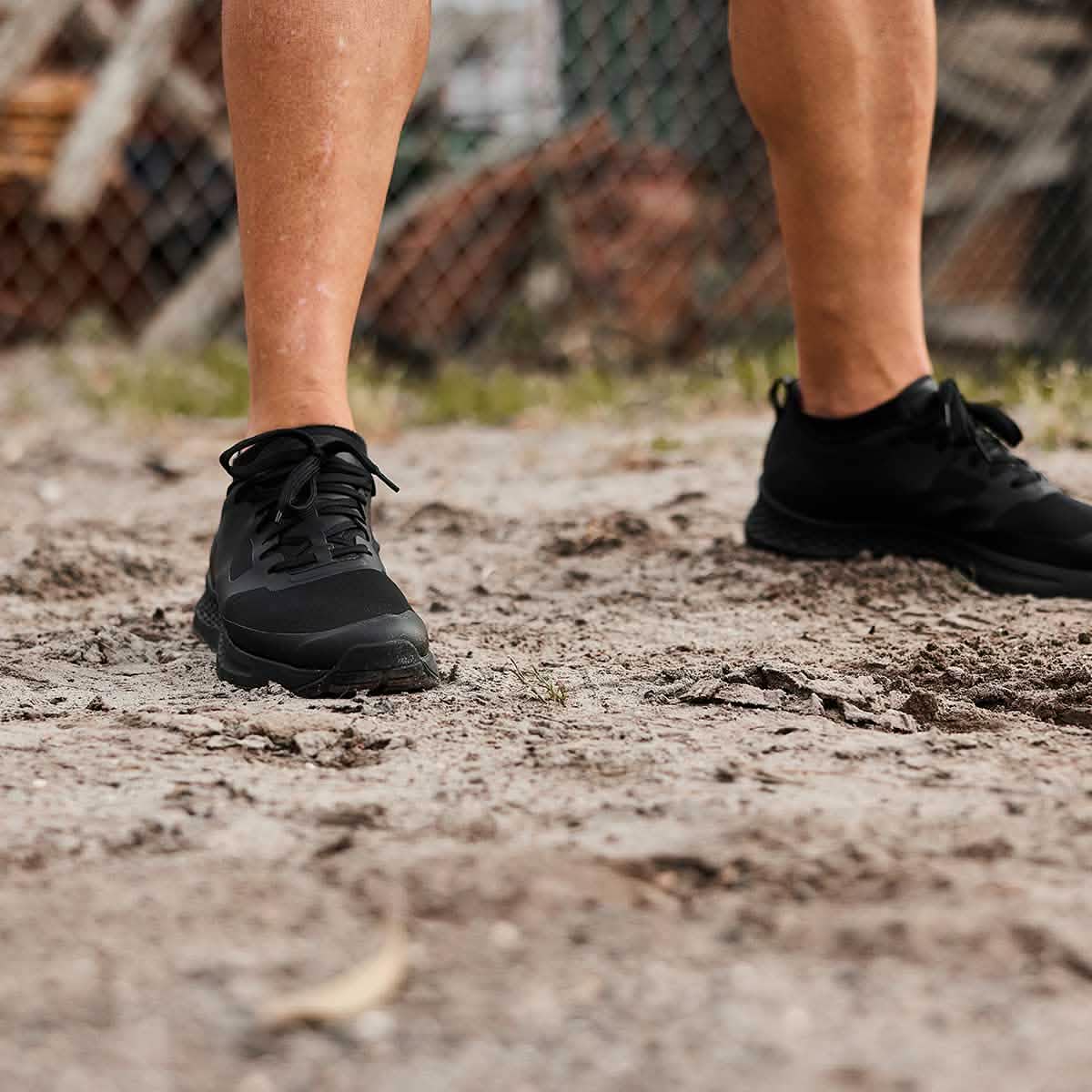 A person wearing GORUCK's Men's Rough Runner - Blackout stands on a dirt path with rocks and grass in the background. The image focuses on the shoes and lower legs, suggesting a Rough Runner event or sporty setting.