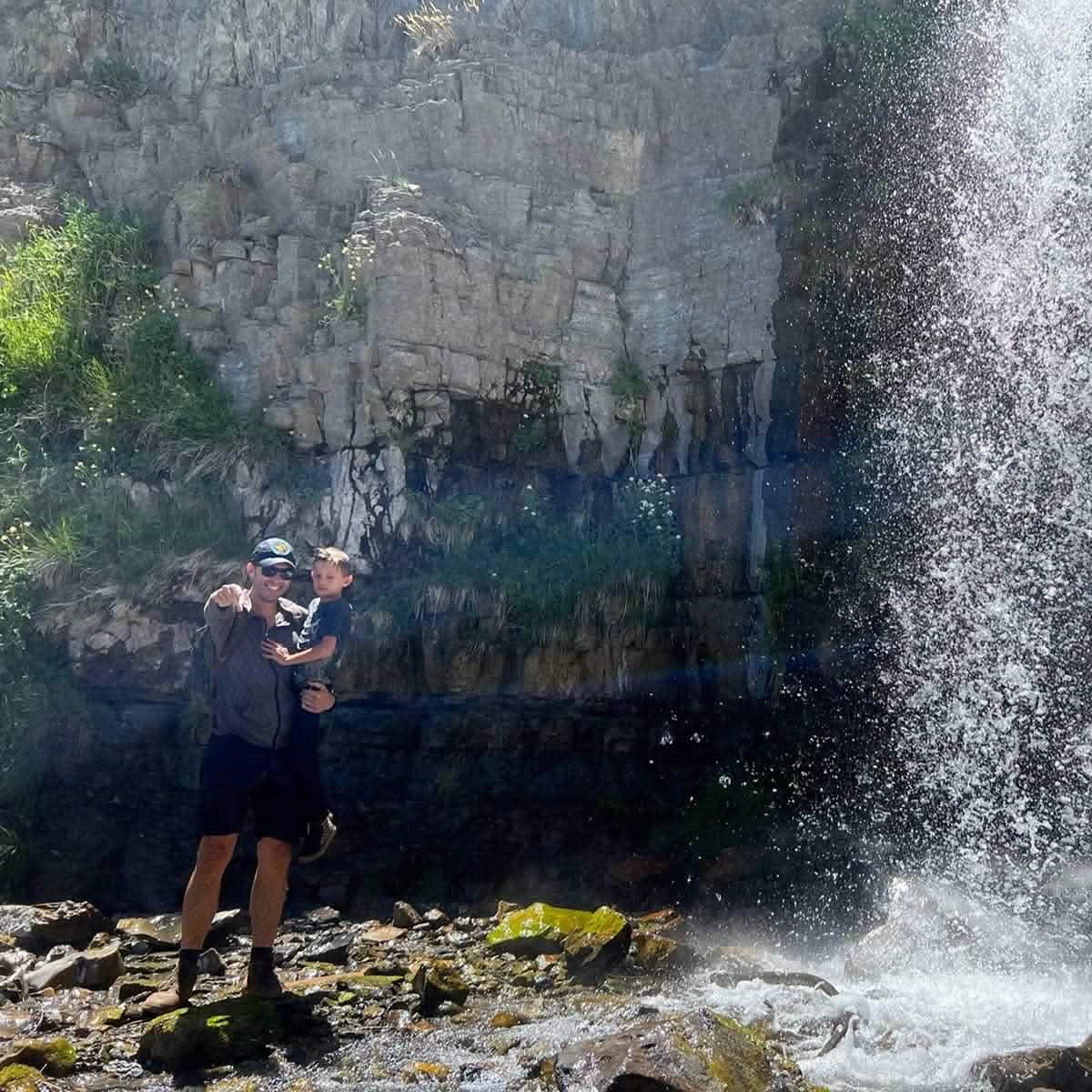 A man wearing GORUCK's Merino Challenge Socks - Crew stands near a waterfall, holding a child as sunlight filters through the spray. Surrounded by rocky terrain and patches of greenery, they enjoy the moment with confidence in their reliable footwear backed by Scars Lifetime Guarantee's promise.
