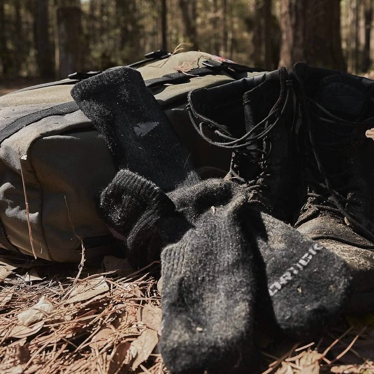 A pair of worn black hiking boots and GORUCK Merino Challenge Socks - Crew rest against a green backpack on a forest floor covered in pine needles. The scene suggests a pause in an outdoor adventure, surrounded by trees. These essentials, made in the USA, promise unmatched reliability and comfort.