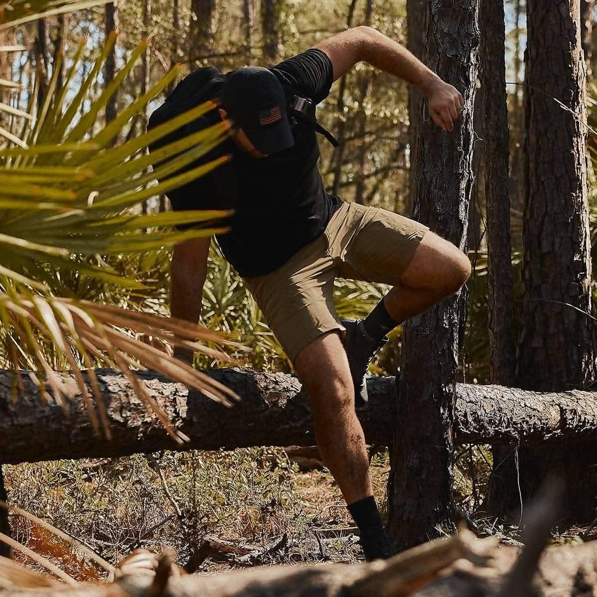 Man in black shirt, cap, and tan shorts climbing over fallen tree in forest, carrying backpack for outdoor rucking