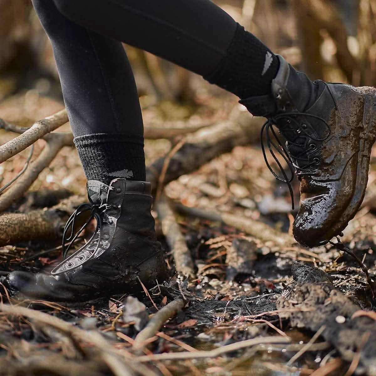 A person wearing black boots and leggings navigates a muddy forest floor scattered with twigs and leaves. The boots, partially covered in mud, are kept comfortable with GORUCK's Merino Challenge Socks - Crew, which promise comfort in every step.