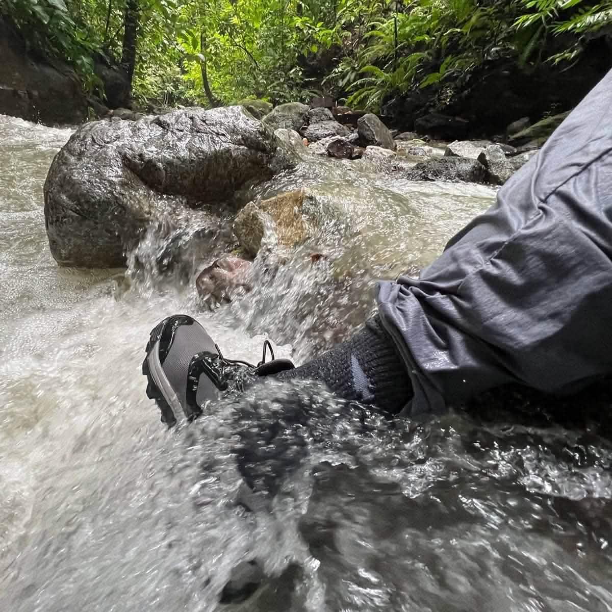 Person wearing hiking shoes and dark pants crossing a flowing rocky mountain stream surrounded by green foliage