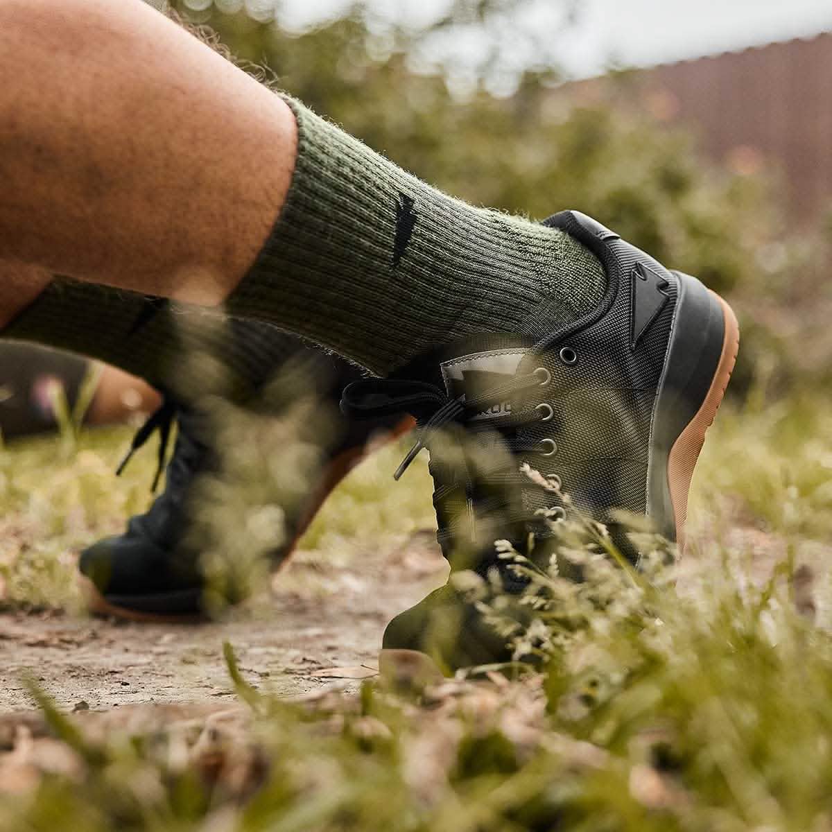 A person sporting GORUCK's Merino Challenge Socks - Crew in green, along with black athletic shoes, is seen walking outdoors on grass. The focus is on the shoes and socks against a softly blurred natural backdrop.