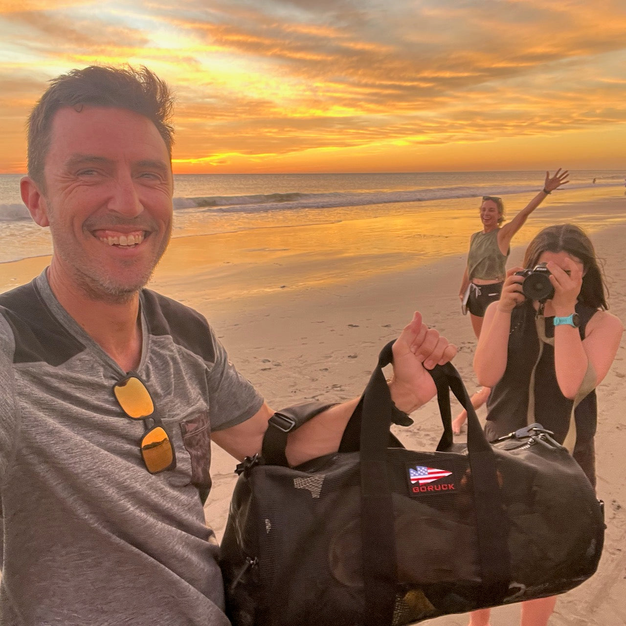 A man smiles on a beach at sunset, holding the Mesh Duffel Bag, while two women stand behind him—one capturing the moment with a photo.