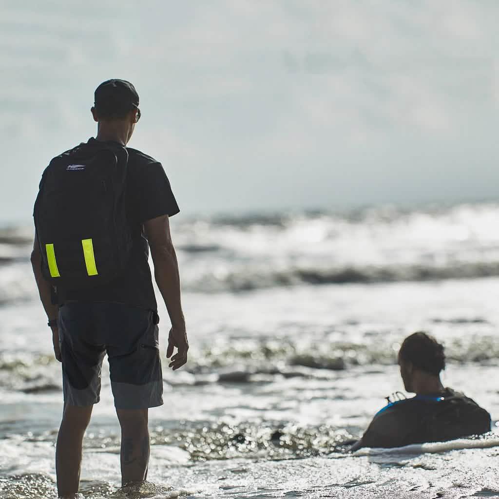 Person with GORUCK backpack in water, rucking on the beach, another person sitting in surf
