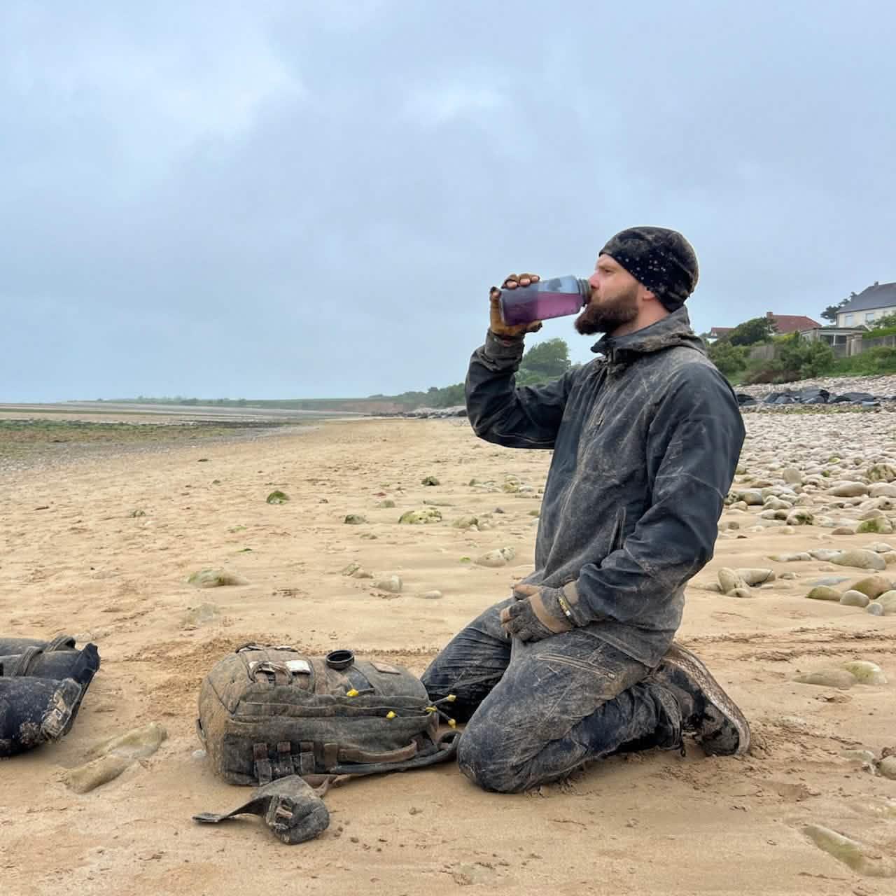 Man in muddy outdoor gear kneeling on a sandy beach, drinking from a Nalgene bottle with GORUCK ruck