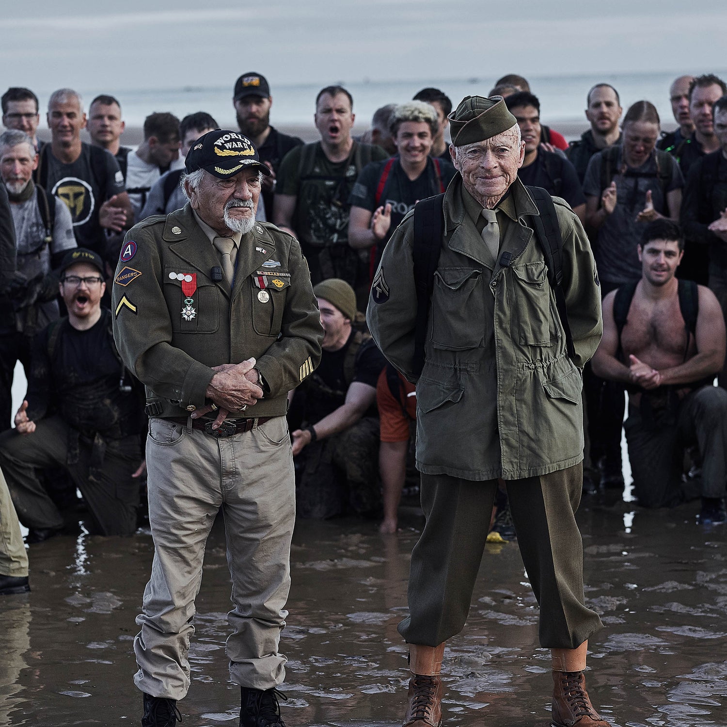 Two elderly veterans in uniform stand on a beach, surrounded by a cheering group of younger people.