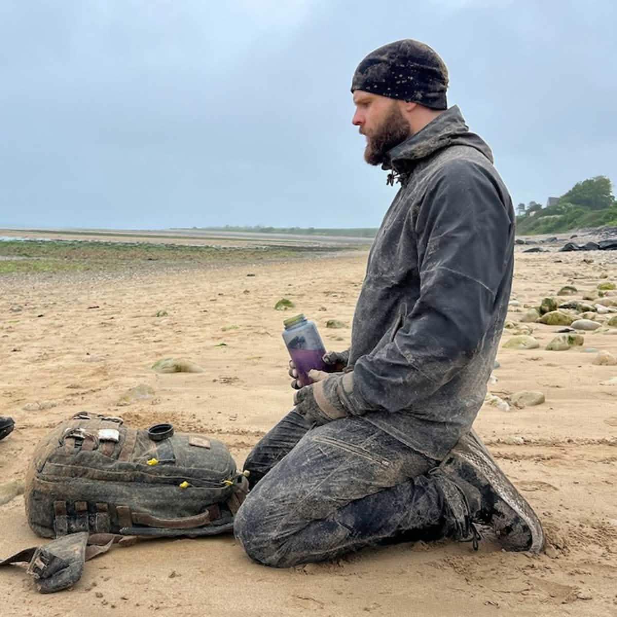 Man in dusty outdoor gear kneeling on a sandy beach holding a water bottle next to a rugged backpack