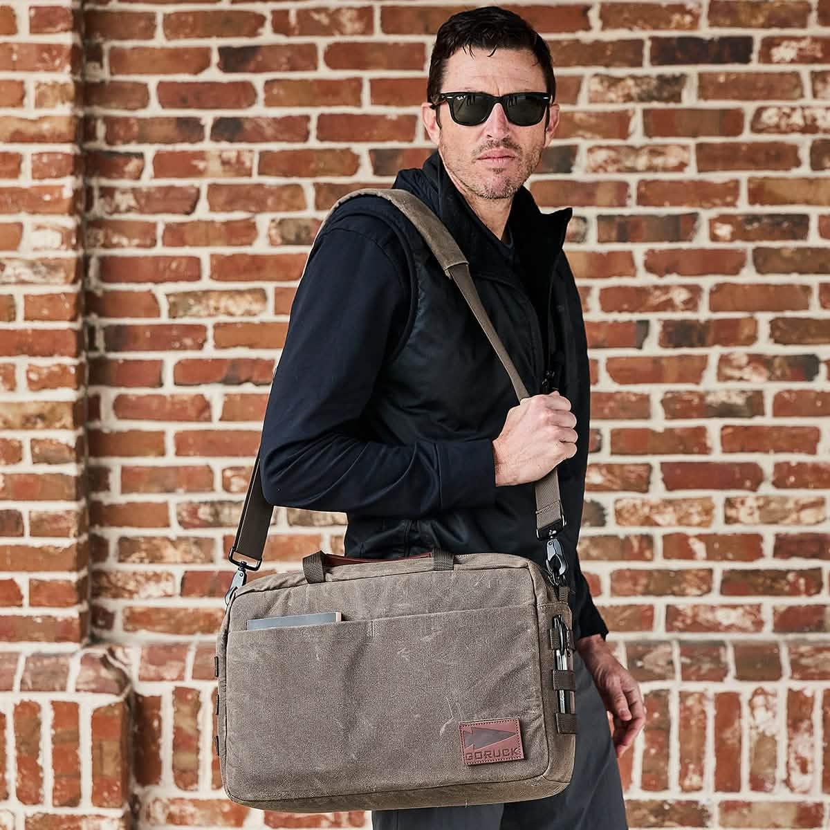 Man wearing sunglasses and black jacket carrying rugged GORUCK Oak canvas messenger bag against brick wall