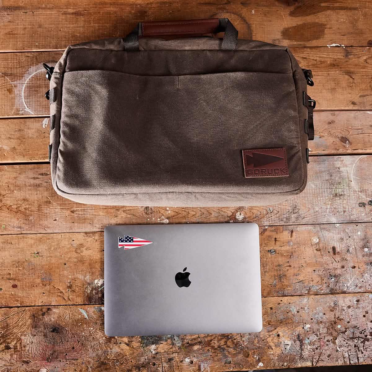 GORUCK rugged brown canvas laptop bag with leather logo patch beside Apple MacBook on wooden surface