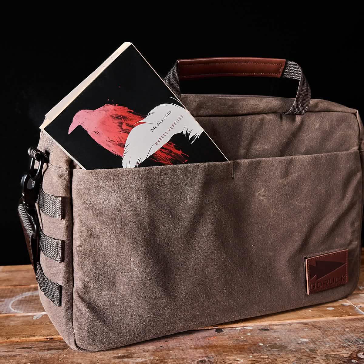 GORUCK durable waxed canvas rucksack with brown leather handle and book in front pocket on wooden surface