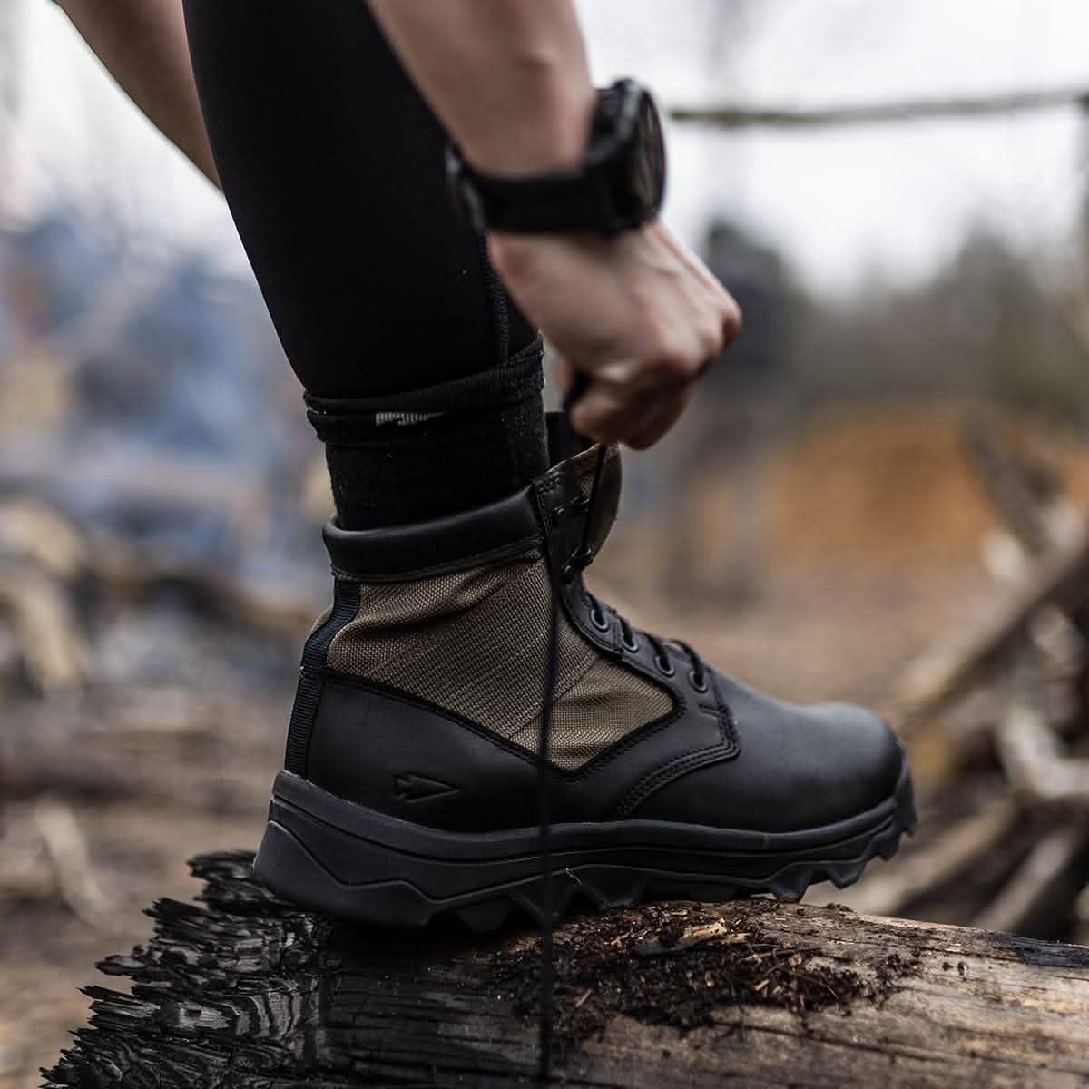 A person wearing a watch is lacing up a rugged MACV-2 - Mid Top - Origins boot by GORUCK, featuring grooved soles, while standing on a charred log. The background is blurred, evoking a forest setting reminiscent of special operations.