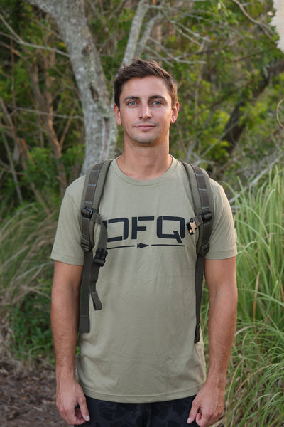 Certainly! Here’s a rewritten version using your product data: A man with short brown hair stands outdoors surrounded by lush greenery, sporting a khaki DFQ Tee - Tri-BlendX and a backpack. The bold “Don’t F*ing Quit” slogan on his shirt gives his adventurous look an extra boost of motivation.