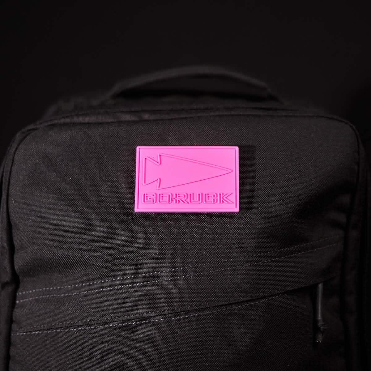 Close-up of black GORUCK backpack featuring a pink patch with embossed arrowhead logo and GORUCK text