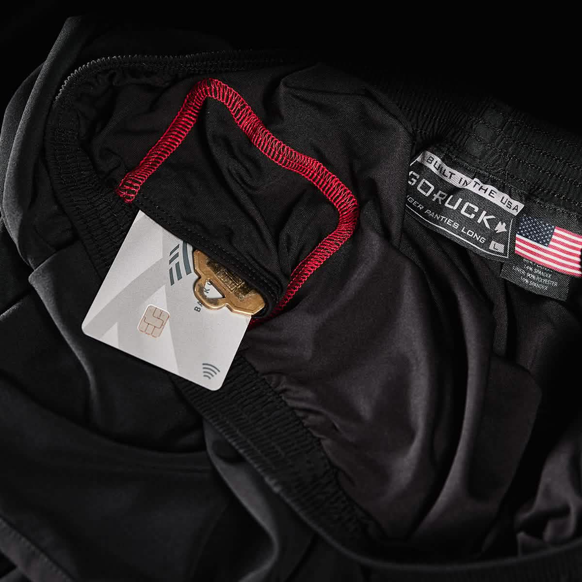 Close-up of the Men's Ranger Panties - ToughStretch by GORUCK, featuring a hidden pocket containing a gold key and credit card. The pocket is outlined in red stitching. An American flag and brand label are visible on the waistband, backed by the Scars Lifetime Guarantee for durability.