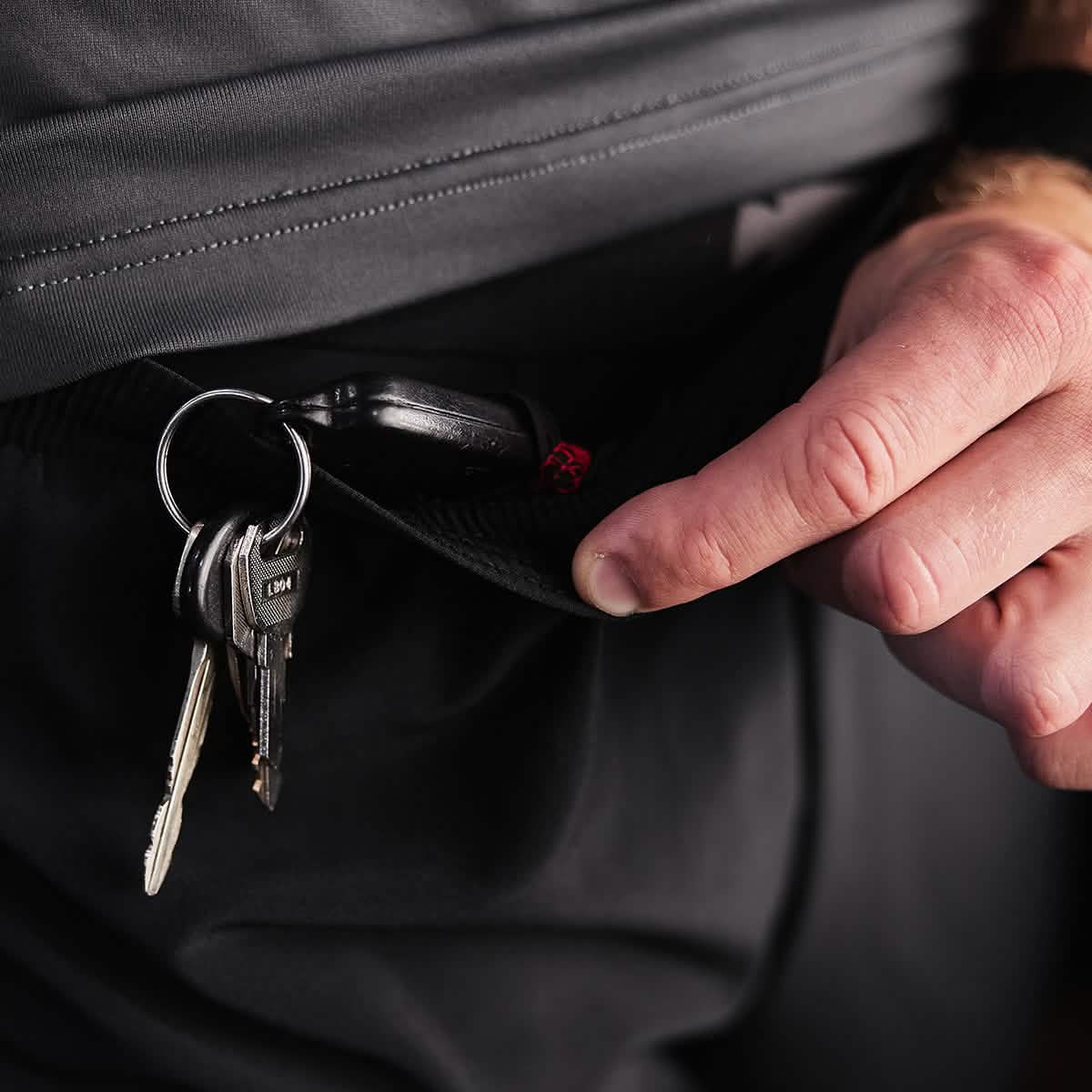 A person grips the waistband of GORUCK's Men’s Ranger Panties - ToughStretch, revealing a set of keys clipped inside. Among the keys are a car key and several silver keys on a ring. The pants, featuring their subtle texture and guaranteed durability through the Scars Lifetime Guarantee, partially obscure the person’s hand.