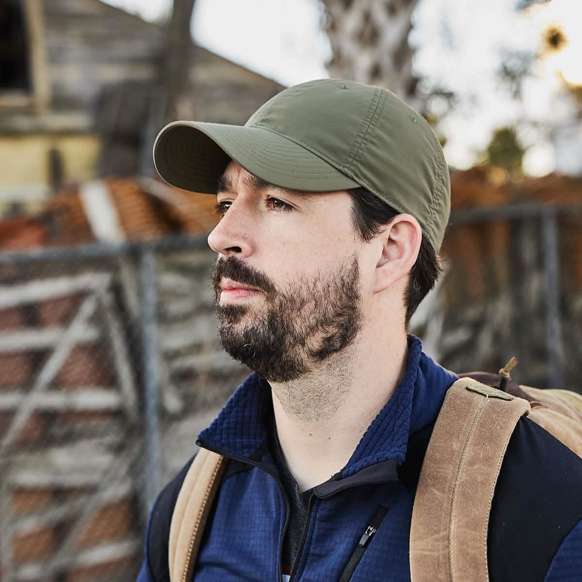 A bearded man, sporting a GORUCK Performance TAC Hat in Slick green and carrying a backpack, stands outdoors. He is dressed in a blue jacket crafted from Sweat-wicking ToughDry® fabric, as he leans casually against a wooden structure with a metal fence visible behind him.