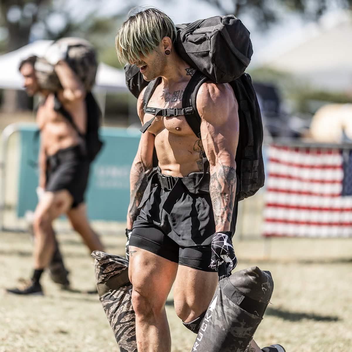 Muscular man in black shorts carrying GORUCK weighted bags during outdoor fitness rucking challenge