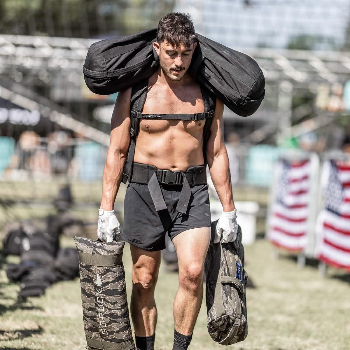 Shirtless man carrying heavy GORUCK sandbags during outdoor rucking event with American flags in background