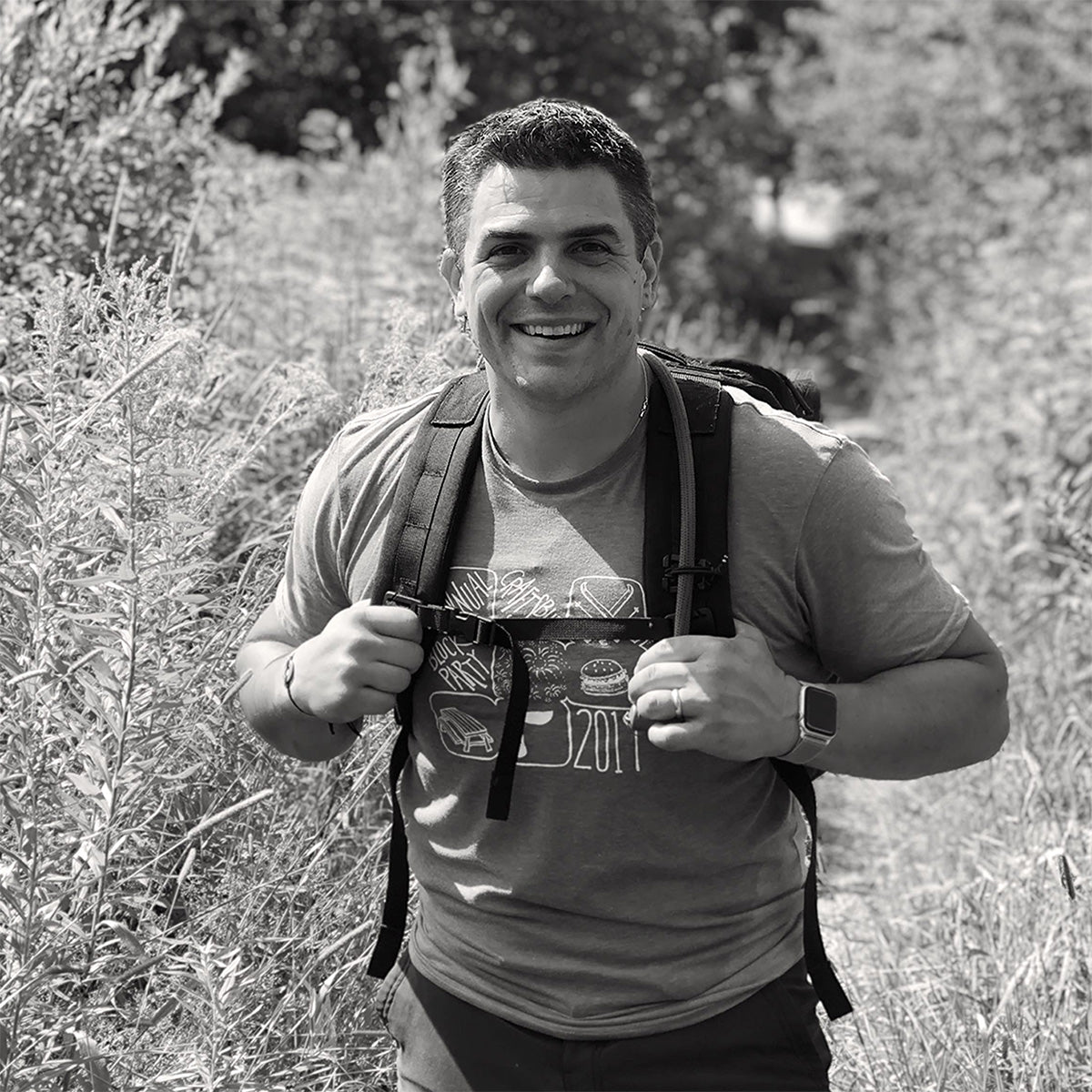 A smiling person with a backpack hikes through tall grass on a sunny day.