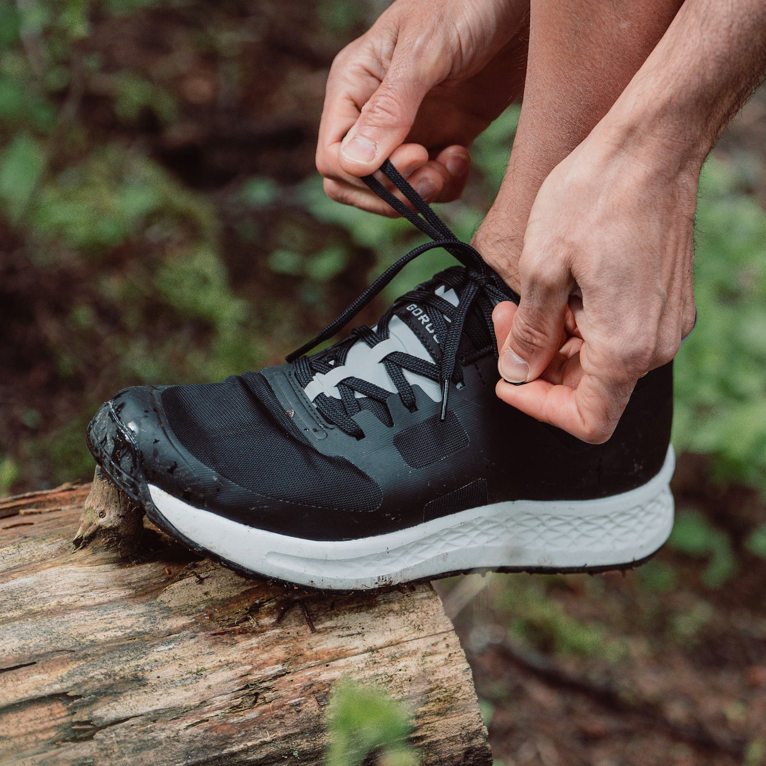 Close-up of hands tying black running shoe laces on a foot resting on a mossy log outdoors.