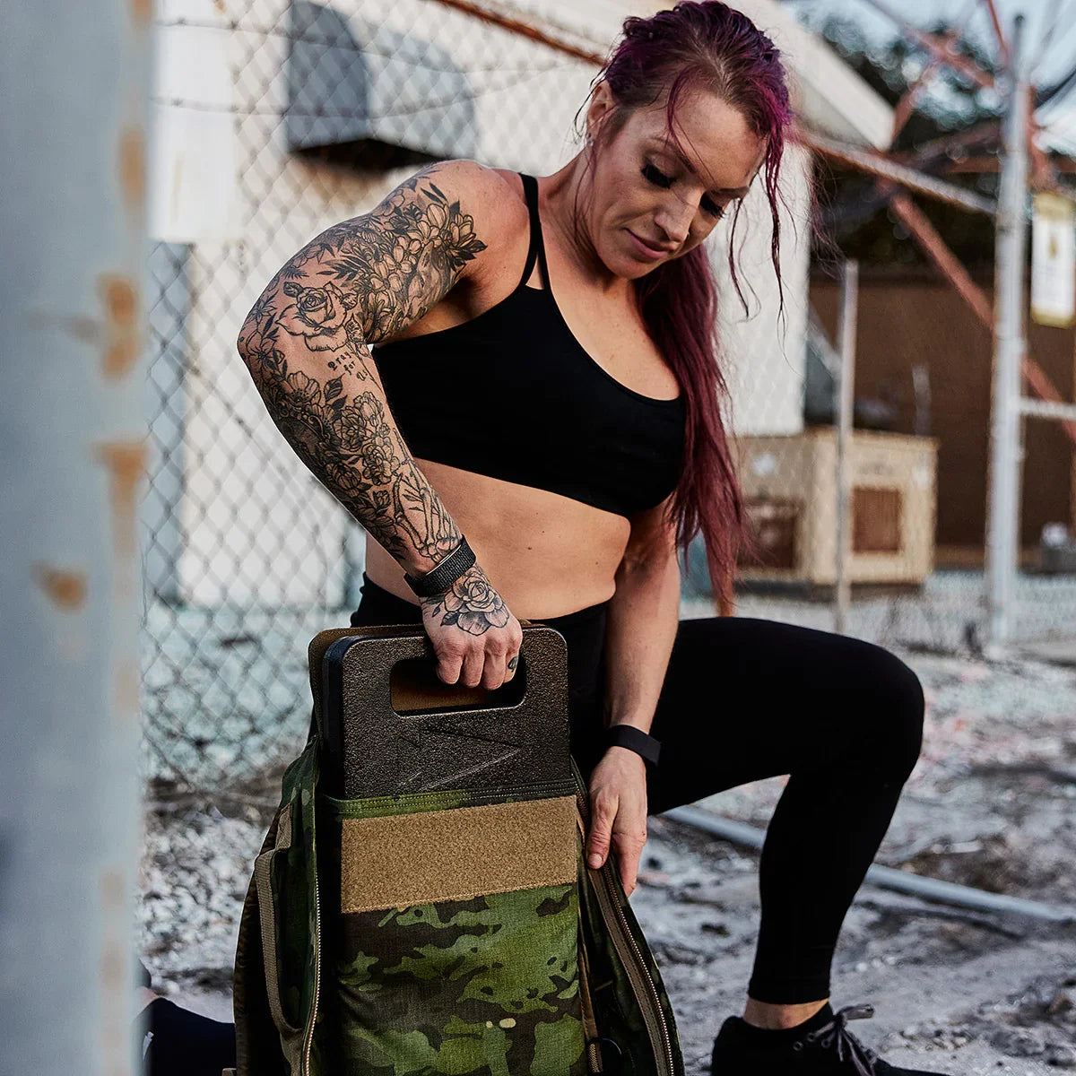 Tattooed woman in black sportswear loading a camouflage GORUCK bag outdoors, fitness training
