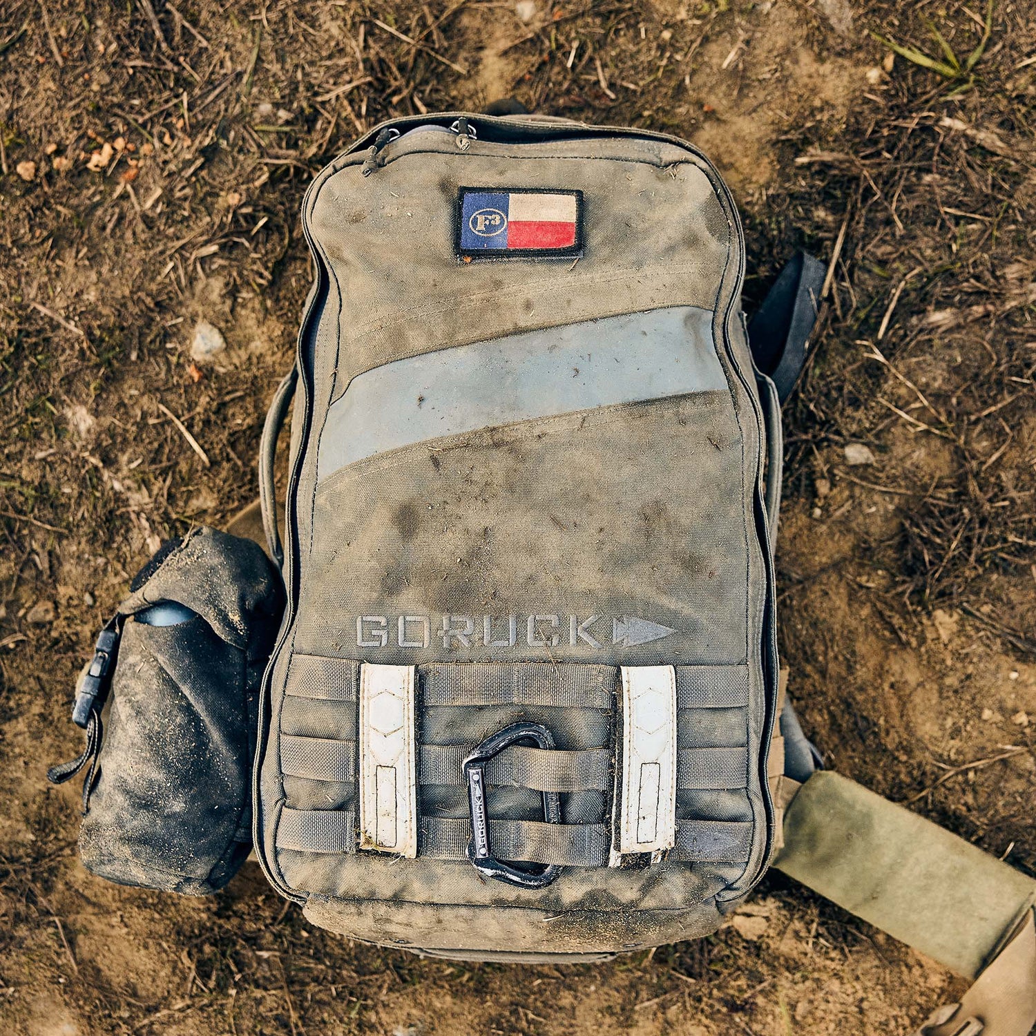 A muddy Rucker 5.0 rucksack rests on dirt, featuring a water bottle attachment and top patch—designed for rucking adventures with built-in durable lumbar support.