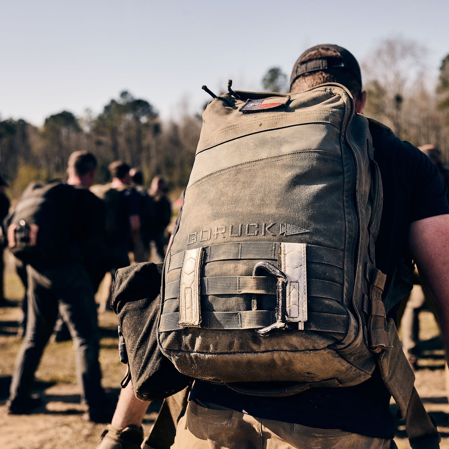 A person stands outdoors in a group on a sunny day, wearing the Rucker 5.0—a large, dusty rucksack known for excellent lumbar support.