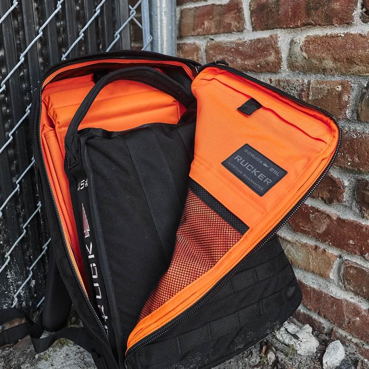 Open GORUCK Rucker 25L backpack with bright orange interior against brick wall and chain-link fence