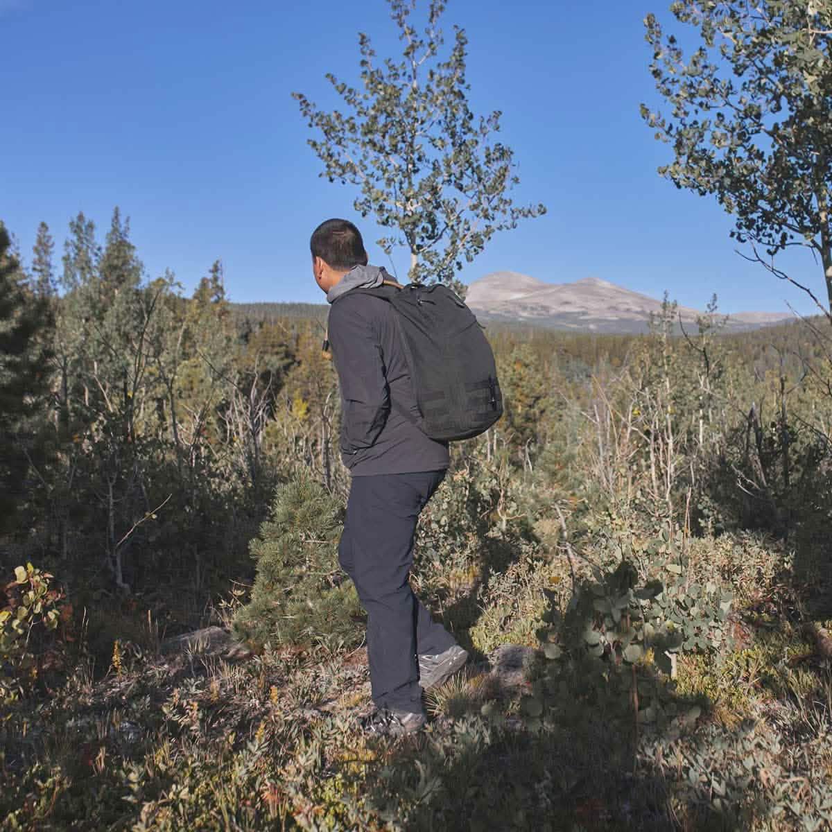 Man wearing black rucking gear with backpack hiking through forested mountainous landscape under clear blue sky