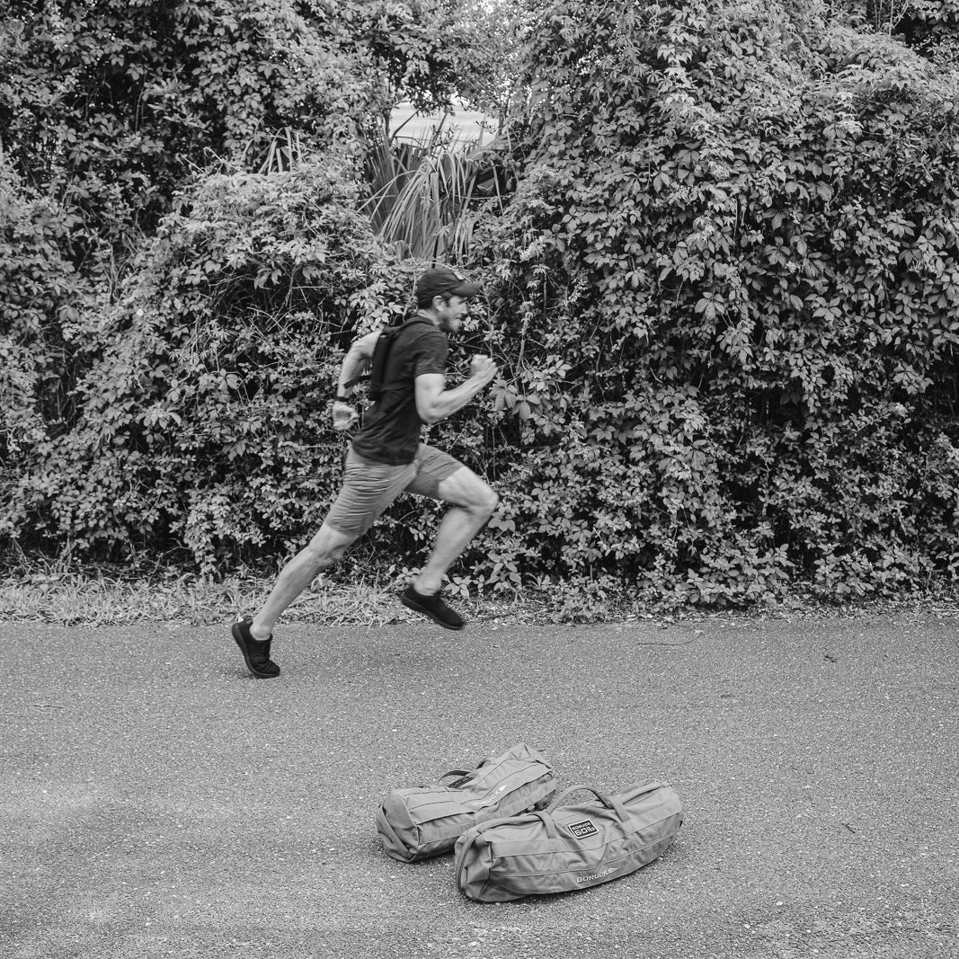 A person runs on a road toward two backpacks, wearing Men’s Simple Shorts - Lightweight ToughDry®, with dense foliage in the background.