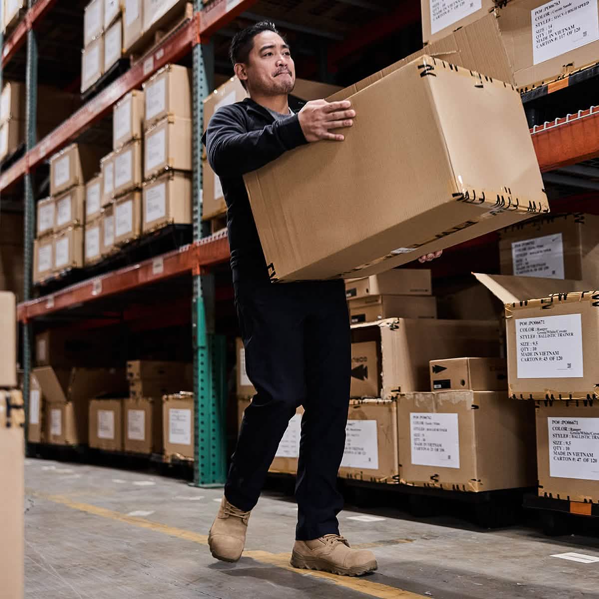 A man carrying a large cardboard box in a warehouse is clad in a dark sweater, pants, and durable MACV-2 Safety Boot - Mid Top by GORUCK that meet Special Forces standards. Shelves filled with similar boxes line the background, reflecting an environment fueled by precision and strength.