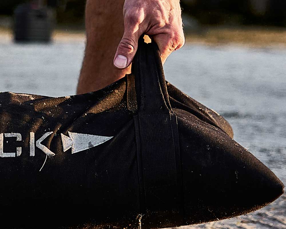 A close-up of a hand gripping the handle of a black sandbag with a white arrow design.