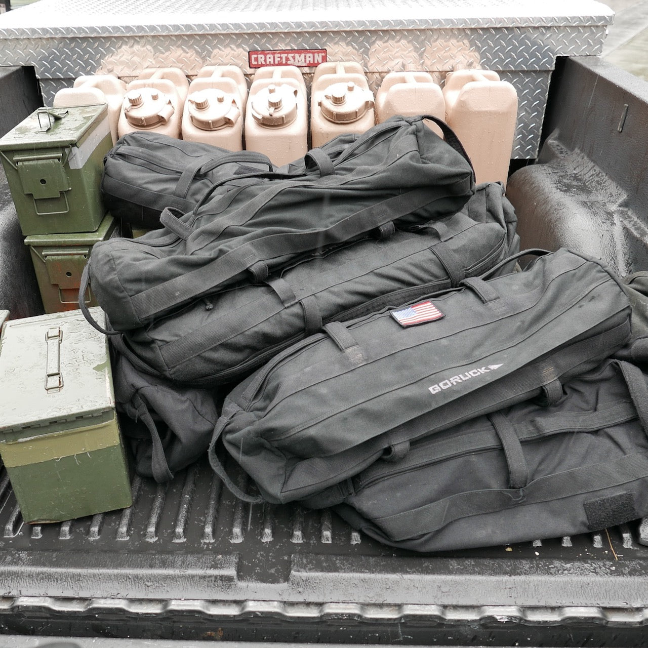Three black duffel bags, two green ammo boxes, tan fuel cans, and a Sandbag Kit (w/ Filler Bag) with lifetime guarantee are in the truck bed.