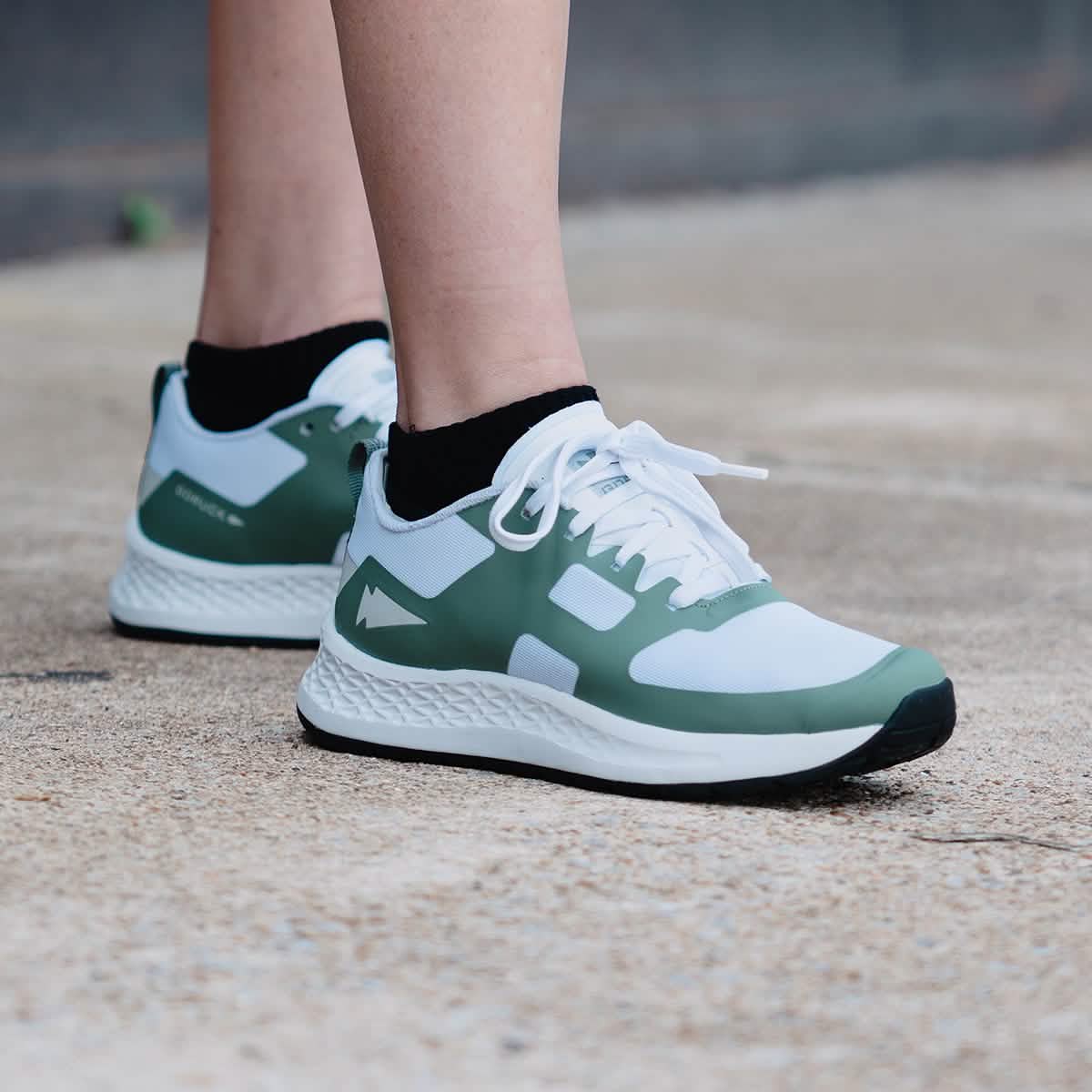Close-up of a person wearing the Women's Rough Runner - Seaspray athletic shoes by GORUCK, featuring a white and green design and black socks on a textured concrete surface. These high mileage running shoes are highlighted by their distinctive logo, layered sole design, and Gradient Density EVA Midsole for enhanced comfort.