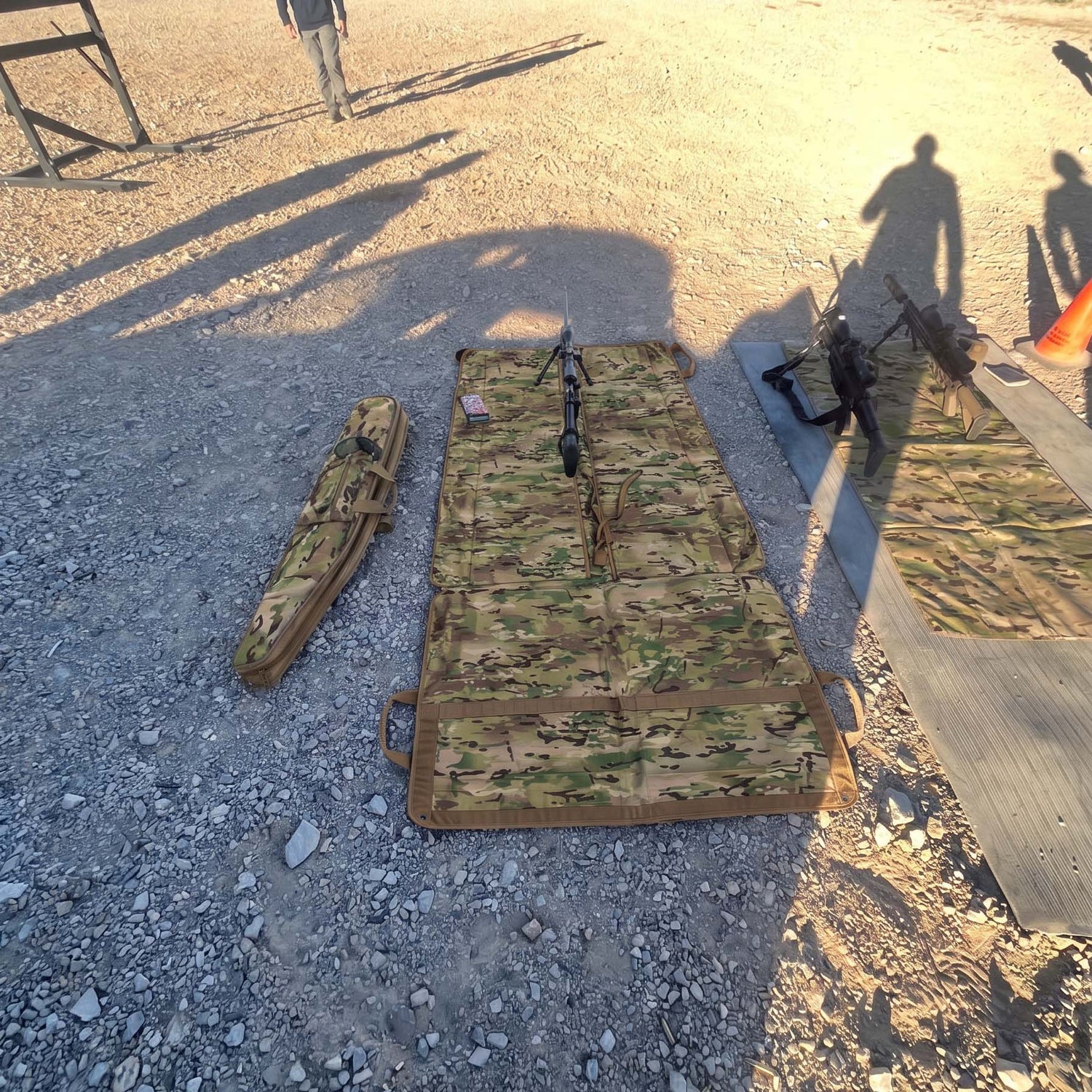 Shooting Mat and rifle on gravel with shadows of people; rifle case and equipment also visible.
