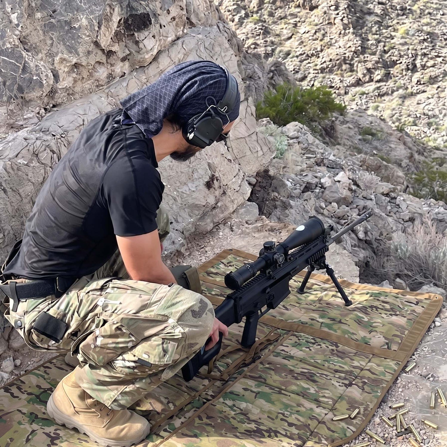 A person kneels on a Shooting Mat with a sniper rifle, surrounded by rocky terrain and wearing camouflage pants.