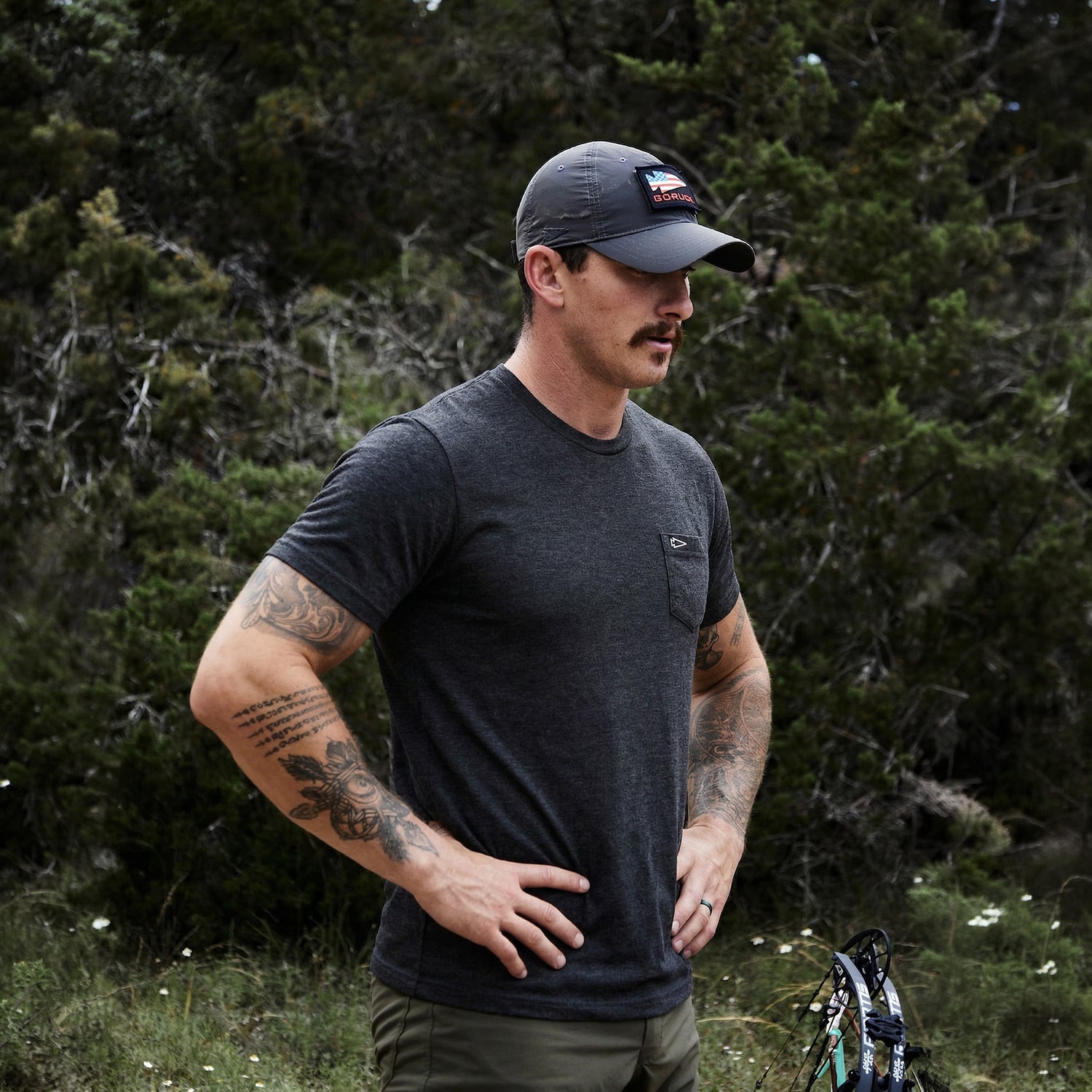 Tattooed man with a mustache stands outdoors among greenery, wearing the Short Sleeve Pocket Tee - Tri-BlendX and a cap.