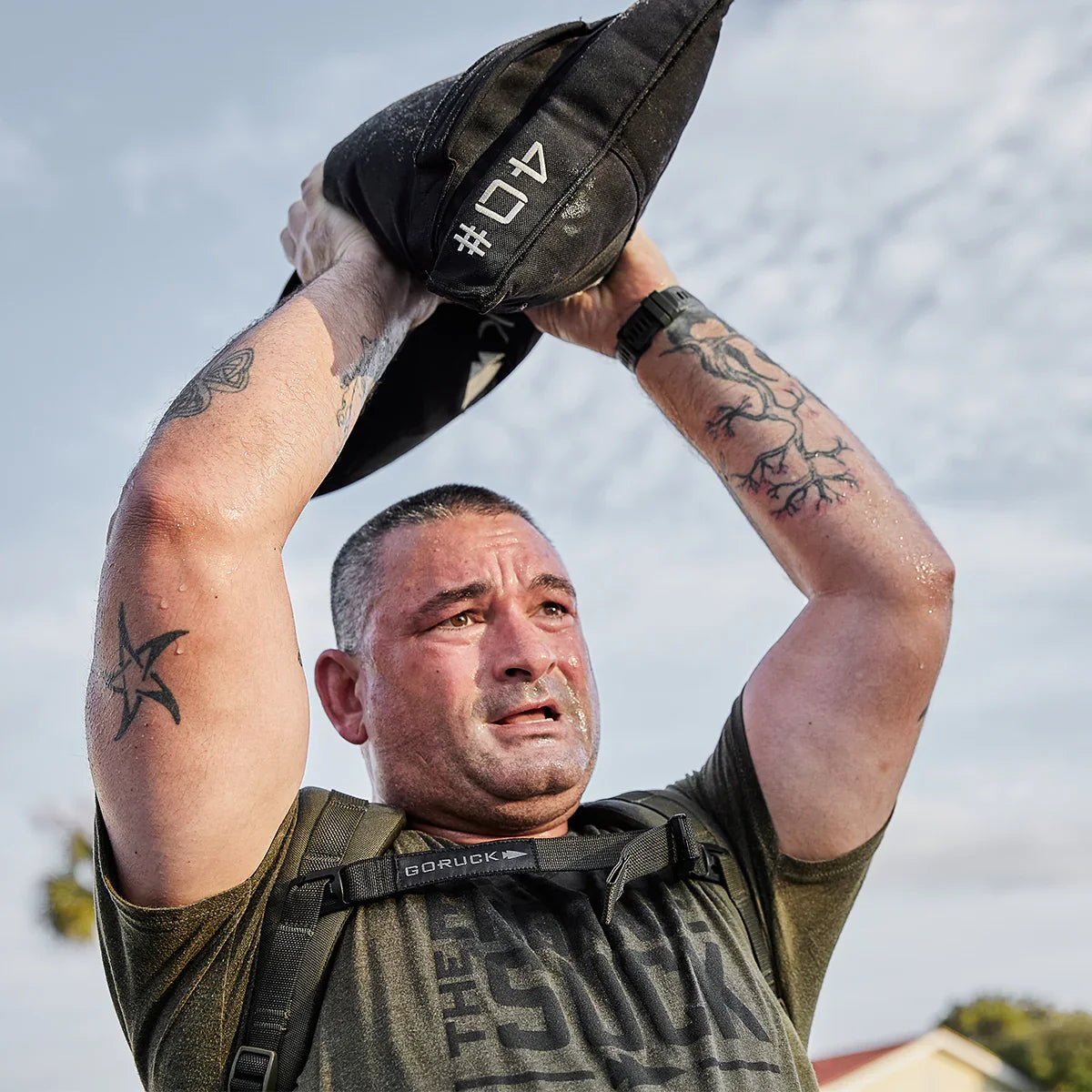 Man lifting GORUCK ruck sandbag outdoors, wearing GORUCK gear, showing tattoos and sweat