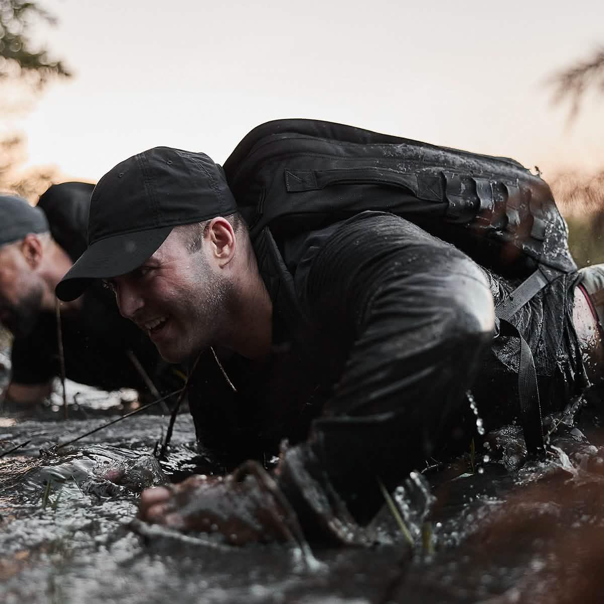 Man wearing black cap and rucking gear crawling through muddy water during outdoor endurance training