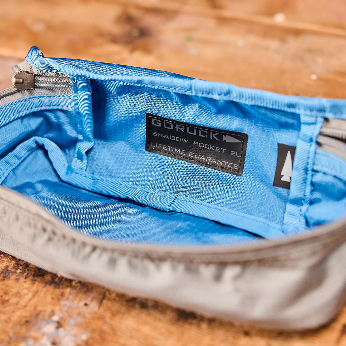 Open gray Shadow Pocket with a blue interior, GORUCK label, and zipper on a wooden surface—ideal for organizing your rucksack. Made of durable 500D Cordura, Shadow Pockets keep your essentials secure and easy to access.