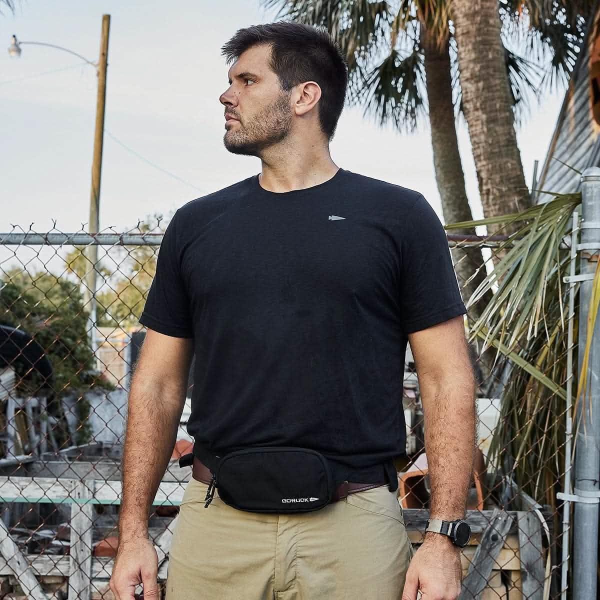 Man wearing GORUCK fanny pack and black t-shirt outdoors near palm tree and fence