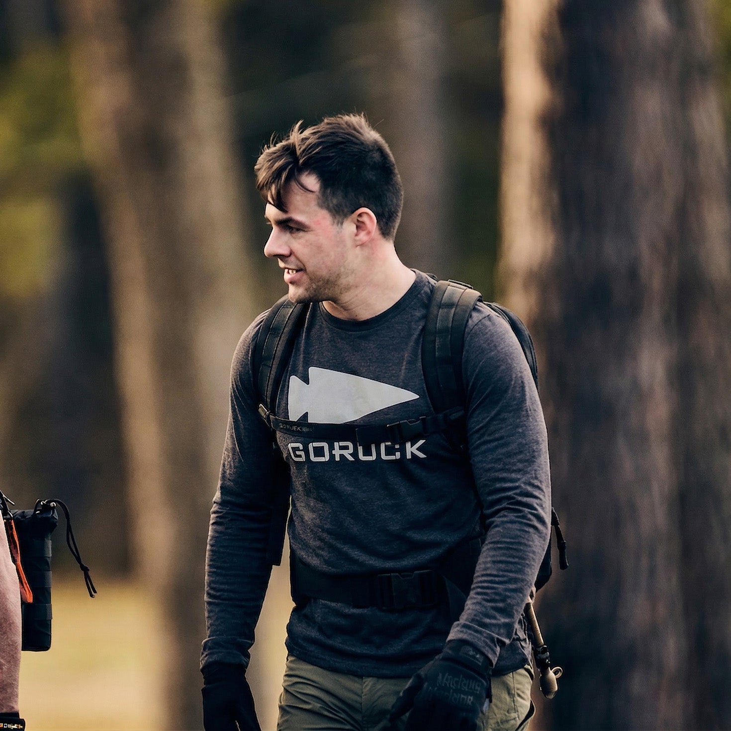 A man in the Men's GORUCK Spearhead Long Sleeve - Tri-BlendX, featuring antimicrobial treatment, smiles outdoors with a backpack in a wooded area.