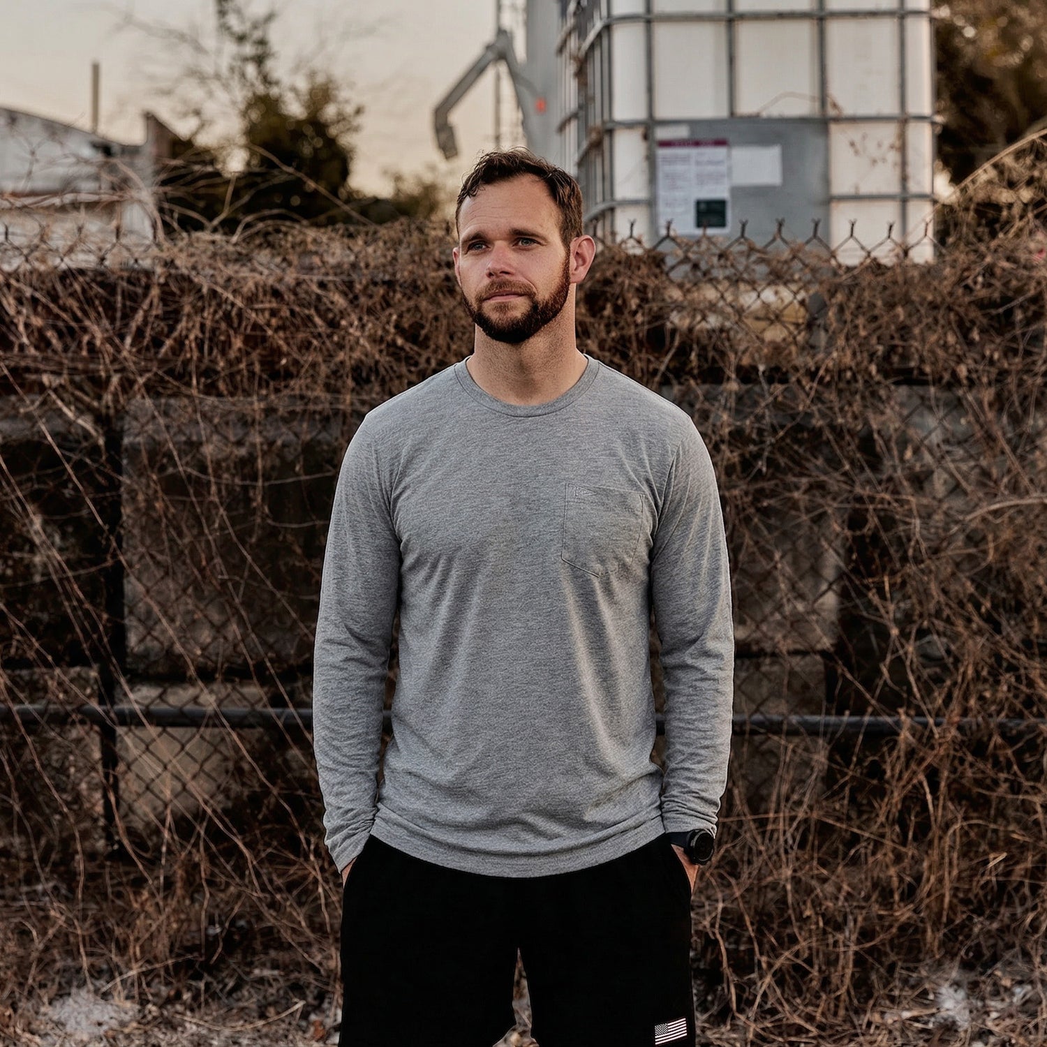 A man in a gray Men's Long Sleeve Pocket Tee - Tri-BlendX stands outdoors in front of a rusted, overgrown chain-link fence.