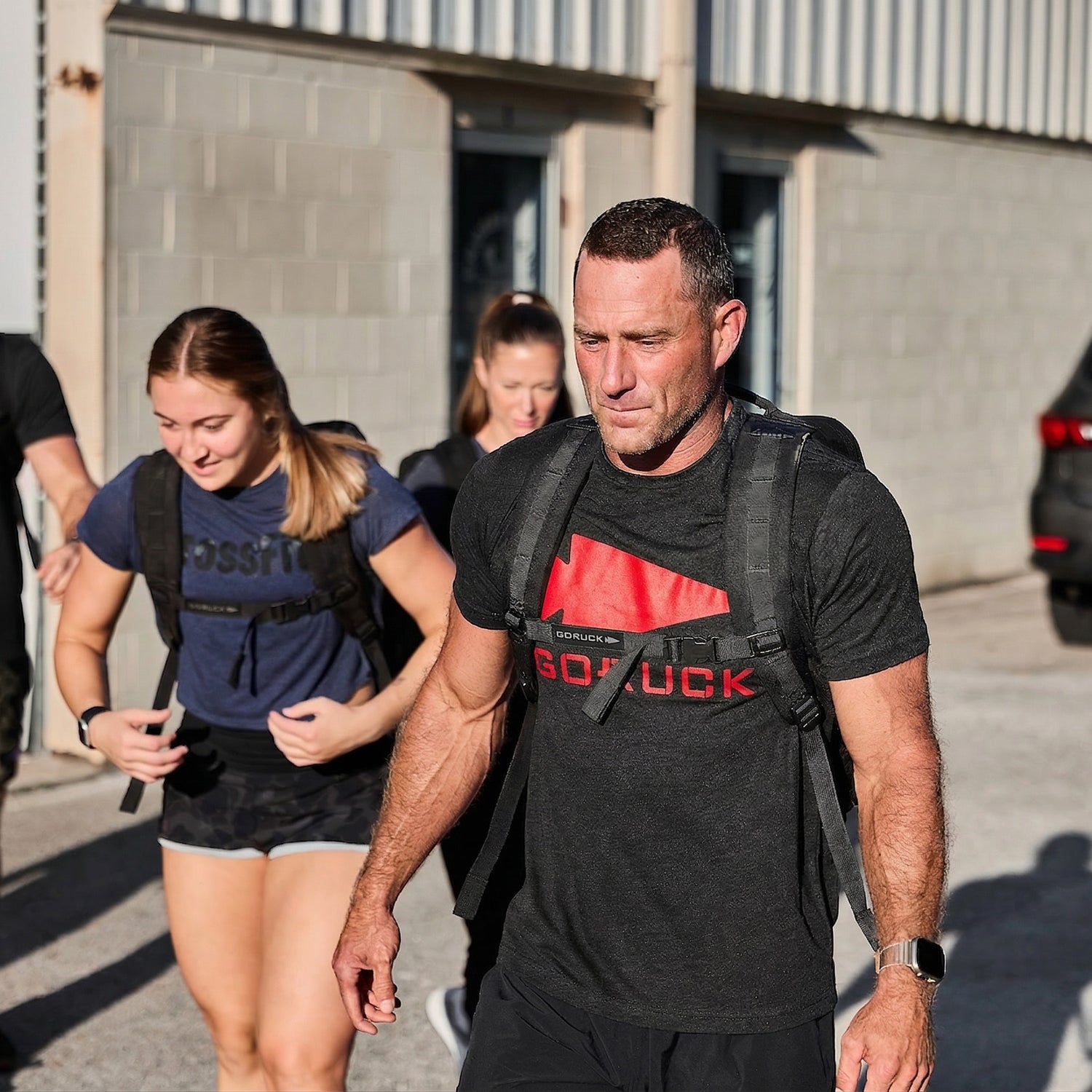 Three people in athletic wear and backpacks walk outdoors in sunlight near a building, each wearing the GORUCK Spearhead Tee - Tri-Blend, featuring the Special Forces–inspired spearhead emblem.