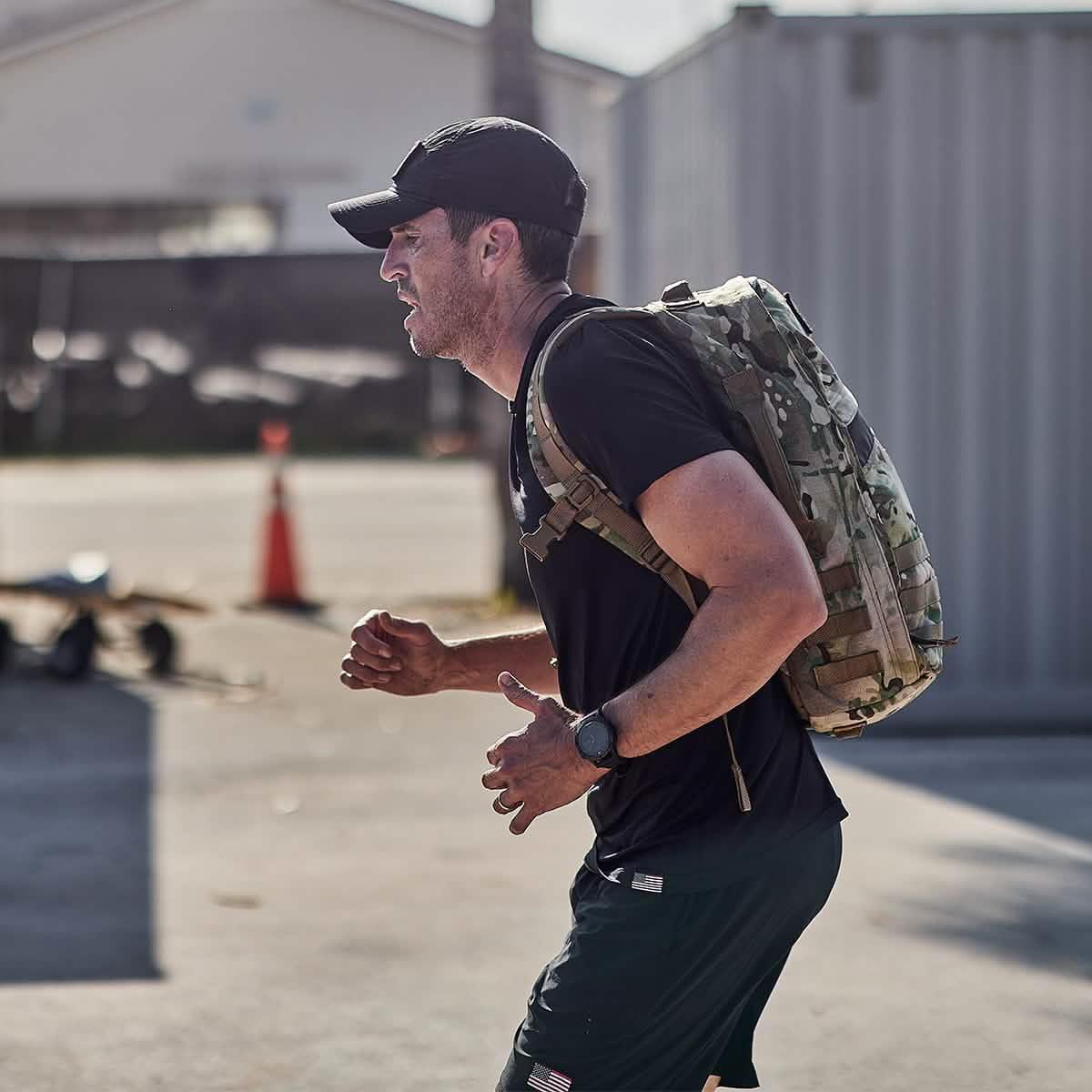 Man running outdoors wearing black athletic wear, cap, and GORUCK camouflage backpack during rucking training
