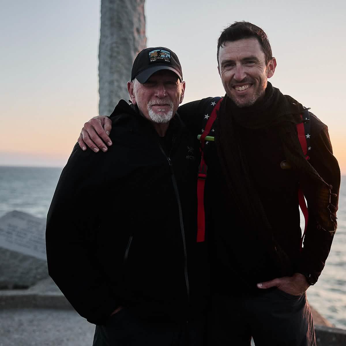Two men in black outdoor gear smiling together by the ocean at sunset, showcasing rugged rucking lifestyle
