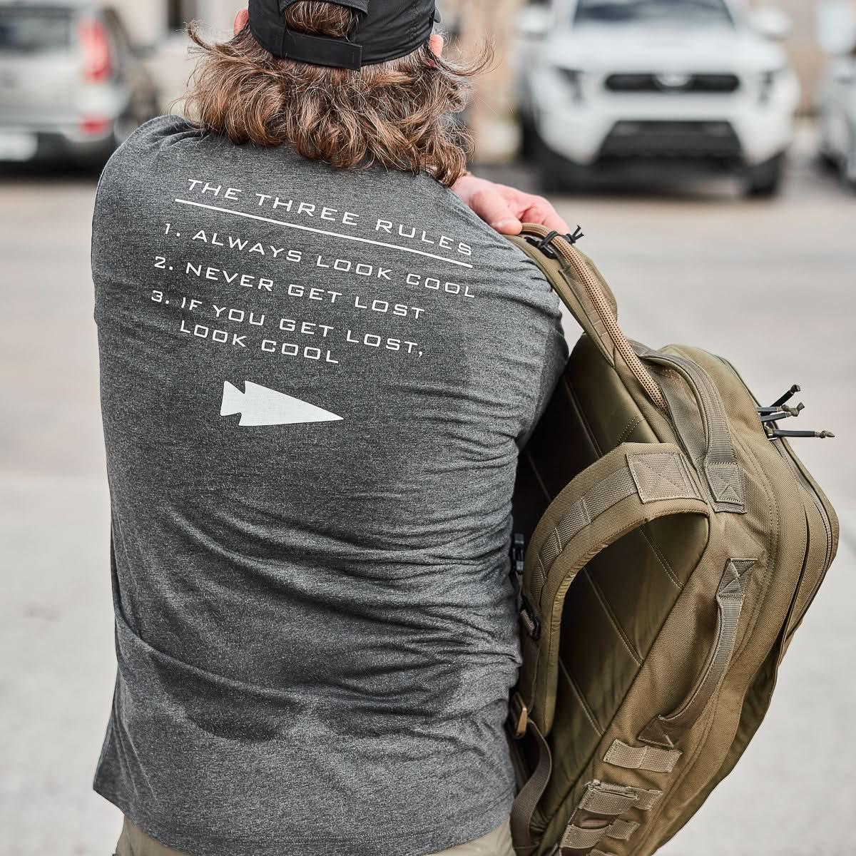 Man wearing charcoal GORUCK The Three Rules tee with white text and carrying a tan tactical rucksack outdoors