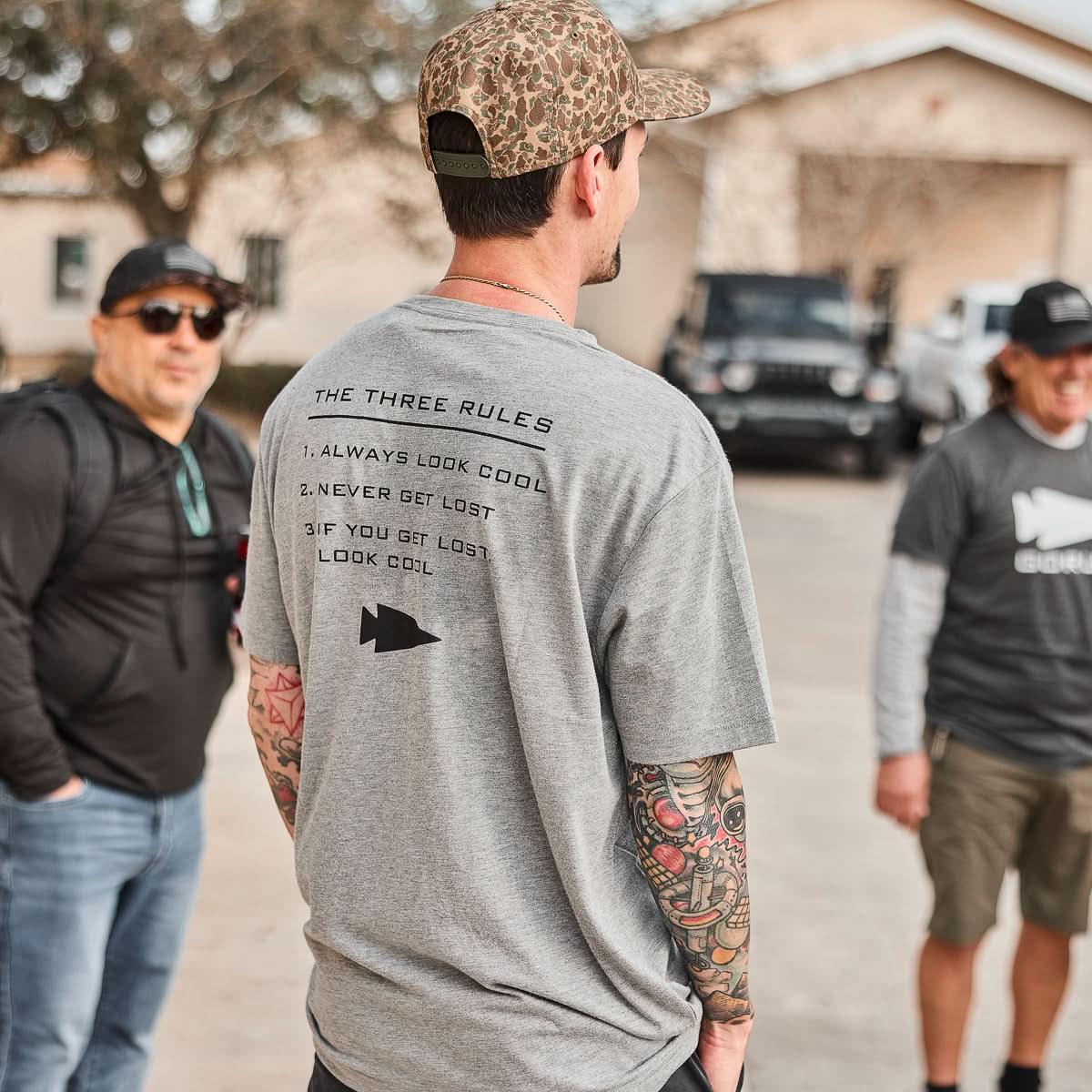 Man wearing grey GORUCK The Three Rules t-shirt with tattooed arms and camouflage hat outdoors